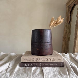 Könnte beinhalten: Eine dunkelbraune Holzvase mit horizontalen Rillen, gefüllt mit getrockneten beigen Gräsern. Die Vase steht auf zwei Büchern, eines mit dem Titel "Curated Style" und das andere mit "Create At Home With Old & New". Die Gegenstände stehen auf einem cremefarbenen Tuch.