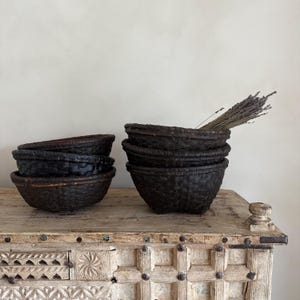 May include: Several dark brown woven baskets are stacked on a light-colored wooden surface. One stack of baskets has a bundle of dried lavender. The baskets have a textured appearance and are of varying sizes. The wooden surface has decorative carvings.