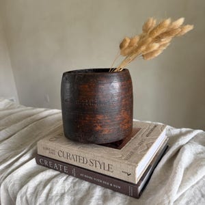 Könnte beinhalten: Eine dunkelbraune, zylindrische Vase mit strukturierter Oberfläche, die getrocknetes, hellbraunes Hasenschwanzgras enthält. Die Vase steht auf zwei Büchern mit den Titeln "CURATED STYLE" und "CREATE". Die Bücher liegen auf einer weißen, strukturierten Oberfläche.