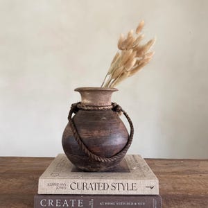 Könnte beinhalten: Eine braune Keramikvase mit einem Seilgriff, die getrocknete beige Blumen enthält. Die Vase steht auf einem Stapel Bücher, darunter eines mit dem Titel "CURATED STYLE". Die Vase hat eine strukturierte Oberfläche und ein einzigartiges, gealtertes Aussehen.