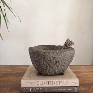 Könnte beinhalten: Eine graue Steinschale mit rauer Textur, die ein kleines Bündel getrockneten Lavendels enthält. Die Schale steht auf zwei Büchern mit den Titeln "CURATED STYLE" und "CREATE" auf einer Holzoberfläche. Ein Pflanzenblatt ist im Hintergrund zu sehen.