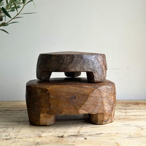 May include: Two stacked, hand-carved wooden stools with a dark brown, weathered finish. Each stool features a thick, circular top supported by three short, sturdy legs. The wood displays visible grain, natural cracks, and a rough, textured surface.