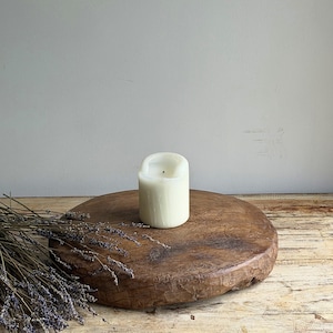 May include: A cylindrical off-white wax candle with a single centered wick, positioned on a thick, circular, weathered wooden board. A bundle of dried purple lavender stems lies across the left side of the textured, dark brown wooden surface.