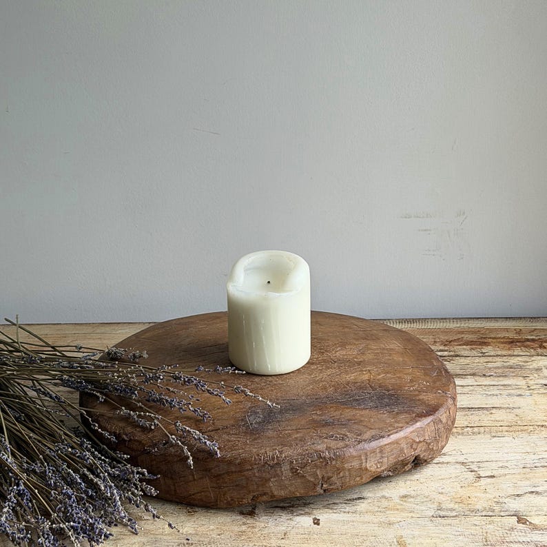 May include: A cylindrical off-white wax candle with a single centered wick, positioned on a thick, circular, weathered wooden board. A bundle of dried purple lavender stems lies across the left side of the textured, dark brown wooden surface.