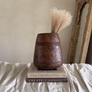 Könnte beinhalten: Eine rustikale, braune Holzvase mit strukturierter Oberfläche, gefüllt mit einem Bündel getrocknetem, beigem Pampasgras. Die Vase steht auf zwei Büchern mit den Titeln "Curated Style" und "Create".