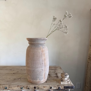 Könnte beinhalten: Eine hohe, verwitterte Holzvase mit breitem Boden und leicht ausgestelltem Rand. Die Vase ist mit zarten weißen Blumen gefüllt. Sie steht auf einem rustikalen Holztisch und erzeugt eine natürliche und Vintage-Ästhetik.