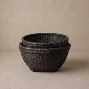 May include: Two dark brown woven baskets, one inside the other. The baskets have a rounded shape and a textured surface. The background is a neutral beige colour.