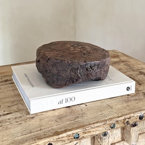 May include: A dark brown, circular wooden block with a rough, weathered texture and visible grain, resting on top of a white hardcover book titled Architectural Digest at 100. The wooden object has a thick, uneven profile and a natural, aged appearance.