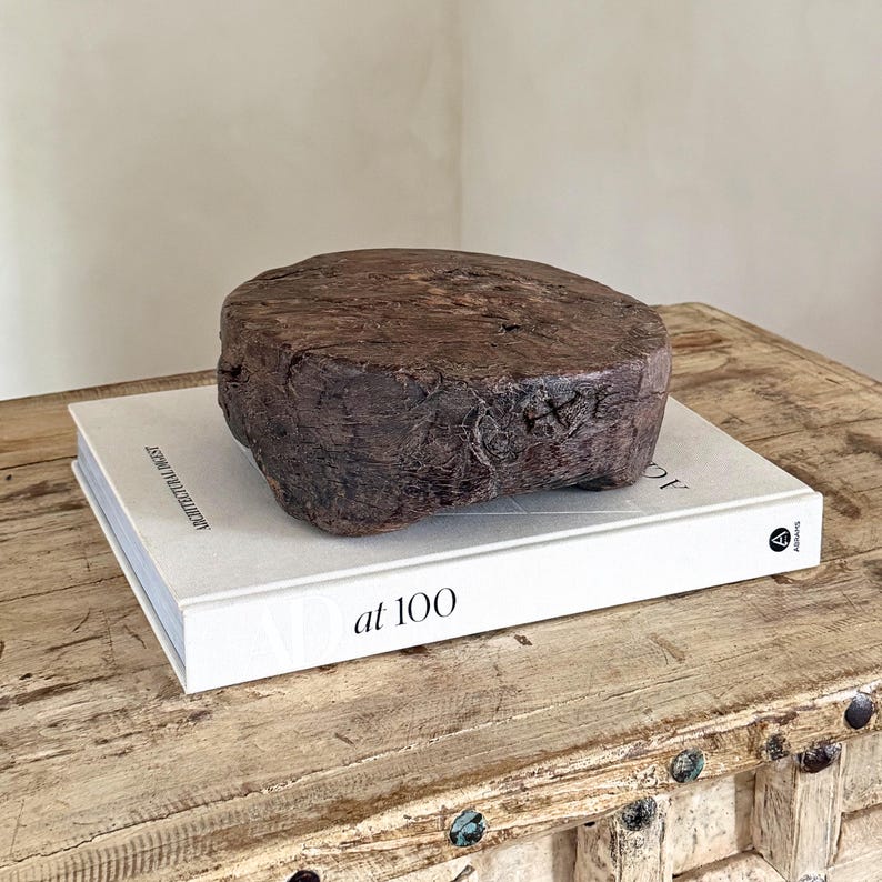 May include: A dark brown, circular wooden block with a rough, weathered texture and visible grain, resting on top of a white hardcover book titled Architectural Digest at 100. The wooden object has a thick, uneven profile and a natural, aged appearance.