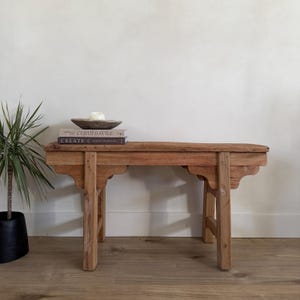 Op de afbeelding: Een rustieke houten bank met een natuurlijke afwerking. De bank heeft een rechthoekige bovenkant en vier stevige poten. Een kleine plant in een zwarte pot en een stapel boeken met een kom en een kaars staan op de bank. De bank is ongeveer 120 cm lang.