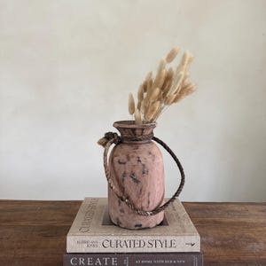 Könnte beinhalten: Eine rustikale, rosafarbene Holzvase mit geflochtenem Seilgriff und einem Bündel getrockneter, beigefarbener Hasenschwanzgras. Die Vase steht auf zwei Büchern mit den Titeln "CURATED STYLE" und "CREATE".