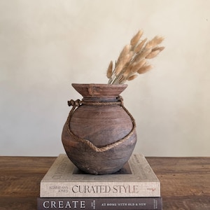 Könnte beinhalten: Eine rustikale, braune Holzvase mit Seildetail, gefüllt mit getrocknetem, beigefarbenem Ziergras. Die Vase steht auf zwei Büchern mit den Titeln "CURATED STYLE" und "CREATE". Der Hintergrund ist ein neutrales Beige. Die Vase ist etwa 20 cm hoch.