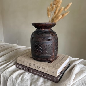 Könnte beinhalten: Eine dunkelbraune, geschnitzte Holzvase mit breiter Öffnung, die getrocknetes Ziergras enthält. Die Vase steht auf zwei Büchern, eines mit dem Titel "Curated Style" und das andere "Create". Die Vase hat ein geometrisches Muster.