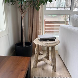 Peut inclure: Un petit tabouret en bois rustique avec un dessus rond et trois pieds. Deux livres sont empilés sur le tabouret. Le tabouret est en bois de couleur claire et a un aspect vieilli. Un pot de plante et une fenêtre sont en arrière-plan.