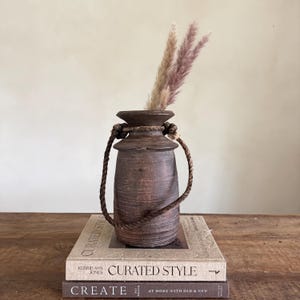 Puede incluir: Un jarrón marrón rústico con un asa de cuerda, que contiene hierba de la pampa seca. El jarrón está sobre dos libros, uno titulado "CURATED STYLE" y el otro "CREATE". El jarrón tiene un aspecto envejecido.