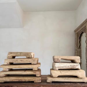 May include: Several stacks of rectangular, light-brown wooden risers with a handle-like cutout on the top. The risers are of varying sizes and are arranged on a dark wooden surface. The background is a neutral-toned wall.