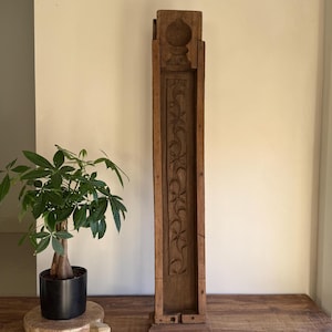Könnte beinhalten: Ein hohes, rechteckiges Holzdekorationsstück mit aufwendigen Blumenschnitzereien. Das gealterte Holz hat einen warmen, braunen Ton. Die Oberseite hat ein abgerundetes Design. Eine kleine Topfpflanze steht in der Nähe.