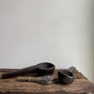 May include: Two dark brown wooden spoons with long handles and deep bowls rest on a weathered wooden surface. A small bundle of dried lavender is tied with twine between the spoons. The spoons are likely for kitchen use.