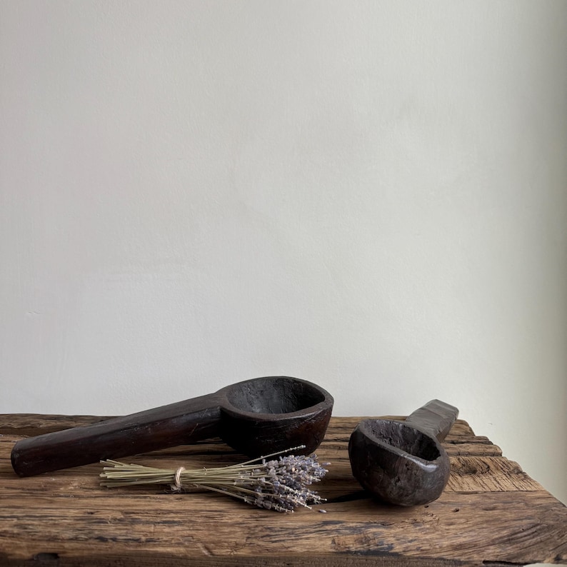 May include: Two dark brown wooden spoons with long handles and deep bowls rest on a weathered wooden surface. A small bundle of dried lavender is tied with twine between the spoons. The spoons are likely for kitchen use.