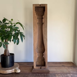Op de afbeelding: Een gebeeldhouwd houten architecturaal element, mogelijk een kolom of baluster, met ingewikkelde details. Het stuk heeft een warme bruine kleur en staat rechtop. Links staat een potplant met groene bladeren.
