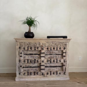 May include: A light-colored wooden cabinet with two doors, adorned with dark metal accents and ring pulls. The cabinet's surface features intricate carvings. A small potted plant and a black rectangular object sit on top.
