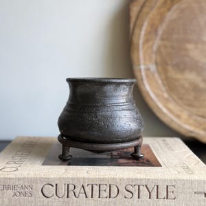 May include: A small, dark gray ceramic pot with a textured surface sits on a dark metal stand with three legs. The pot has a rounded body and a slightly flared rim. The pot is on a book with the words "Curated Style".