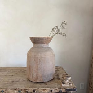 Könnte beinhalten: Eine große, verwitterte Holzvase mit breitem Boden und ausgestelltem Oberteil. Die Vase ist mit zarten, getrockneten weißen Blumen gefüllt. Sie steht auf einem rustikalen Holztisch und erzeugt eine warme, natürliche Ästhetik.