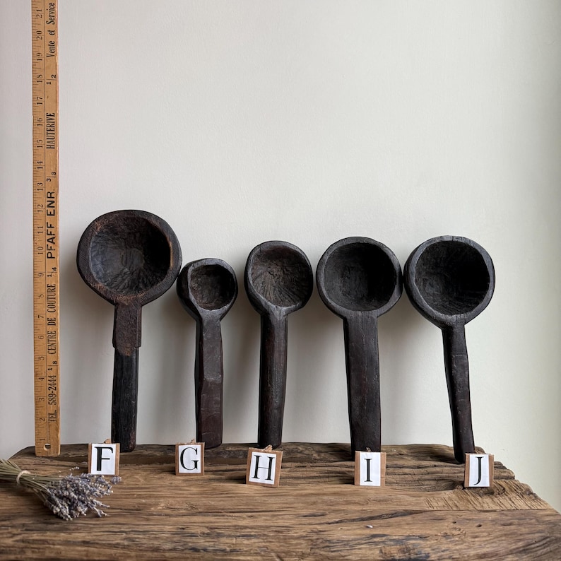 May include: Five dark wooden spoons of varying sizes are displayed on a wooden surface. The spoons have deep bowls and long handles. A measuring ruler is visible on the left. The spoons are labeled F, G, H, I, and J.