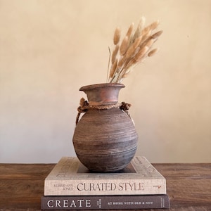 Könnte beinhalten: Eine braune Keramikvase mit Seildetails, gefüllt mit getrocknetem, beigem Ziergras. Die Vase steht auf zwei Büchern, eines mit dem Titel "CURATED STYLE" und das andere "CREATE". Der Hintergrund ist ein neutrales Beige.
