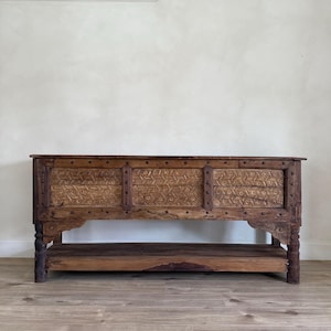May include: A long, rectangular wooden cabinet with intricate carved geometric patterns on the front panels. The cabinet has a lower shelf and is supported by four turned legs. The wood has a warm, natural finish.