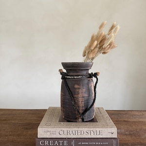 Könnte beinhalten: Eine dunkelbraune, rustikale Vase mit Seilgriff und einem Bündel getrockneter beiger Blumen. Die Vase steht auf zwei Büchern mit den Titeln "Curated Style" und "Create".