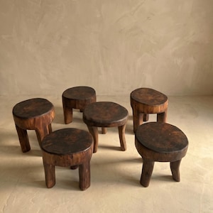 Puede incluir: Seis pequeños taburetes de madera rústicos con un acabado marrón oscuro. Cada taburete tiene un asiento redondo y tres o cuatro patas robustas. Los taburetes están dispuestos en grupo, mostrando su veta natural de la madera y formas únicas. Los taburetes miden unos 30 cm de altura.