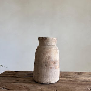 Könnte beinhalten: Eine rustikale, hellfarbene Holzvase mit breitem Boden und schmalerem Hals. Die Vase hat eine zylindrische Form und steht auf einer verwitterten Holzoberfläche. Die Holzmaserung ist sichtbar und trägt zur natürlichen Ästhetik bei. Die Vase ist leer.