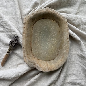 Könnte beinhalten: Eine rustikale, beige Steinschale mit ovalem Innenraum, auf einem zerknitterten, hellgrauen Stoff. Ein kleines Bündel getrockneten Lavendels, mit braunem Papier gebunden, liegt links von der Schale. Die Schale hat eine strukturierte Oberfläche.
