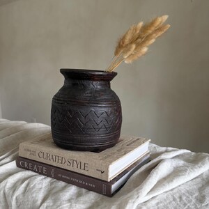 Könnte beinhalten: Eine dunkelbraune, strukturierte Vase mit Zickzackmuster, die getrocknete, hellbraune Gräser enthält. Die Vase steht auf zwei Büchern mit den Titeln "Curated Style" und "Create". Der Hintergrund ist ein neutrales Off-White.