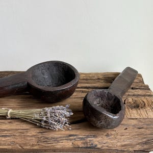 May include: Two dark brown, hand-carved wooden scoops with long handles rest on a weathered wooden surface. A bundle of dried lavender is tied with twine. The scoops have a rustic, antique appearance.