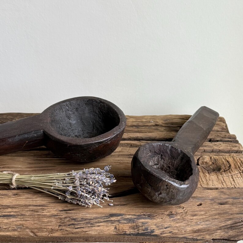 May include: Two dark brown, hand-carved wooden scoops with long handles rest on a weathered wooden surface. A bundle of dried lavender is tied with twine. The scoops have a rustic, antique appearance.