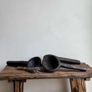 May include: Three dark wooden scoops with long handles rest on a rustic wooden table. The scoops have a deep, rounded bowl shape. A small bundle of dried lavender sits near one of the scoops. The wood has a textured, aged appearance.
