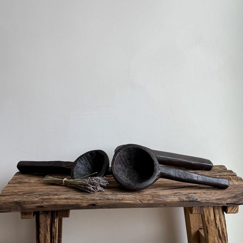 May include: Three dark wooden scoops with long handles rest on a rustic wooden table. The scoops have a deep, rounded bowl shape. A small bundle of dried lavender sits near one of the scoops. The wood has a textured, aged appearance.