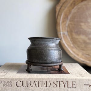 May include: A small, dark gray ceramic pot with a rounded body and flared rim, resting on a dark metal stand with three short legs. The pot sits atop a book with the words "CURATED STYLE" on the cover.