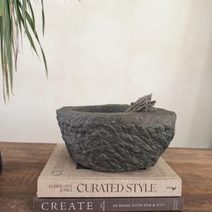 Puede incluir: Un mortero de piedra gris oscuro con una superficie rugosa y texturizada. Un pequeño manojo de lavanda seca se encuentra dentro del mortero. El mortero descansa sobre dos libros con los títulos "CURATED STYLE" y "CREATE".