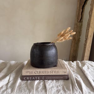 Könnte beinhalten: Eine dunkelbraune, runde Holzvase mit breiter Öffnung, die ein Bündel getrockneter, hellbrauner Gräser enthält. Die Vase steht auf zwei Büchern mit den Titeln "Curated Style" und "Create". Die Anordnung befindet sich auf einer cremefarbenen Leinenoberfläche.