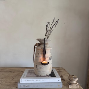 Puede incluir: Un jarrón rústico blanquecino con un portavelas de metal oscuro y una vela de té encendida. El jarrón está adornado con un asa de cuerda y lleno de lavanda seca. El jarrón está sobre una pila de libros en una mesa de madera.