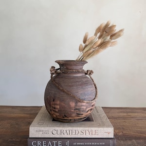 Könnte beinhalten: Eine rustikale, braune Holzvase mit Seilgriffen, gefüllt mit getrockneten, beigen Blumen. Die Vase steht auf zwei Büchern, darunter "Curated Style". Die Vase hat eine runde Form und eine strukturierte Oberfläche, was ihren Vintage-Charme unterstreicht.