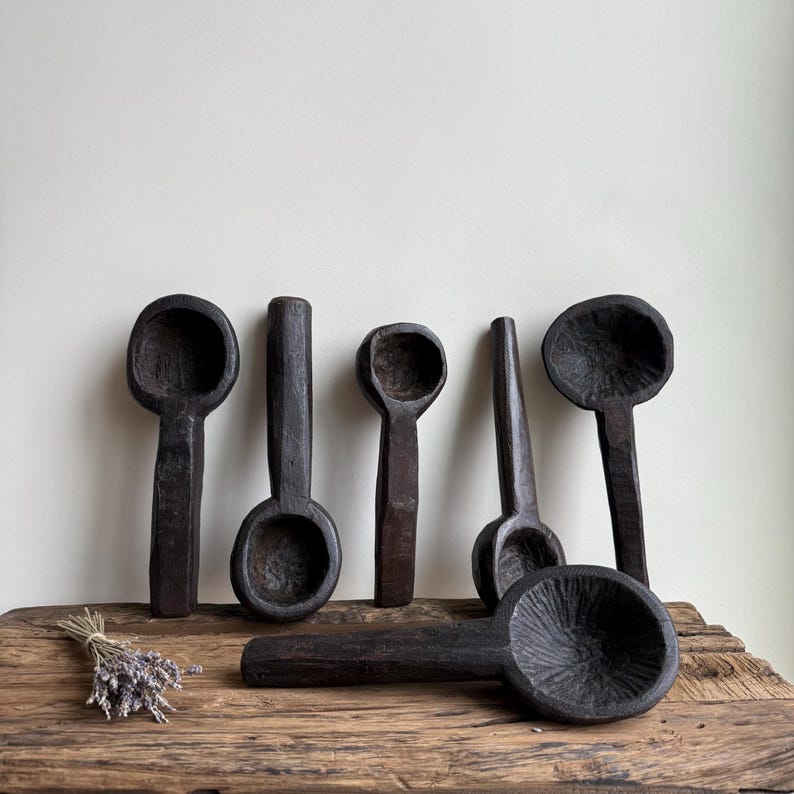 May include: A collection of six dark brown wooden spoons of varying sizes and shapes. The spoons are arranged on a rustic wooden surface, with a small bundle of dried lavender to the left. The spoons have a hand-carved appearance.