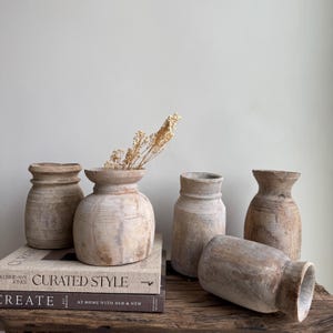 May include: A collection of five rustic wooden vases in varying sizes and shapes. One vase holds dried flowers. The vases are arranged on a wooden surface with books that read "CURATED STYLE" and "CREATE". The vases have a natural, weathered appearance.