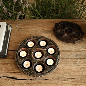 May include: A circular dark brown wooden candle holder with seven recessed circular wells, each containing a small white tea light candle. One candle holder is lit with seven glowing flames, while a second empty holder sits nearby.
