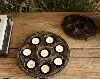 May include: A circular dark brown wooden candle holder with seven recessed circular wells, each containing a small white tea light candle. One candle holder is lit with seven glowing flames, while a second empty holder sits nearby.