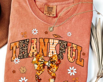 May include: A coral-colored Comfort Colors sweatshirt with the word "THANKFUL" in a fall-themed design. The letters feature leopard print, plaid, and floral patterns. A gold necklace and a brown hat are also visible.
