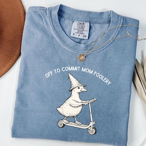 May include: A light blue t-shirt with the text "OFF TO COMMIT MOM FOOLERY" and a cartoon duck wearing a wizard hat and riding a scooter. A gold necklace is draped across the shirt.